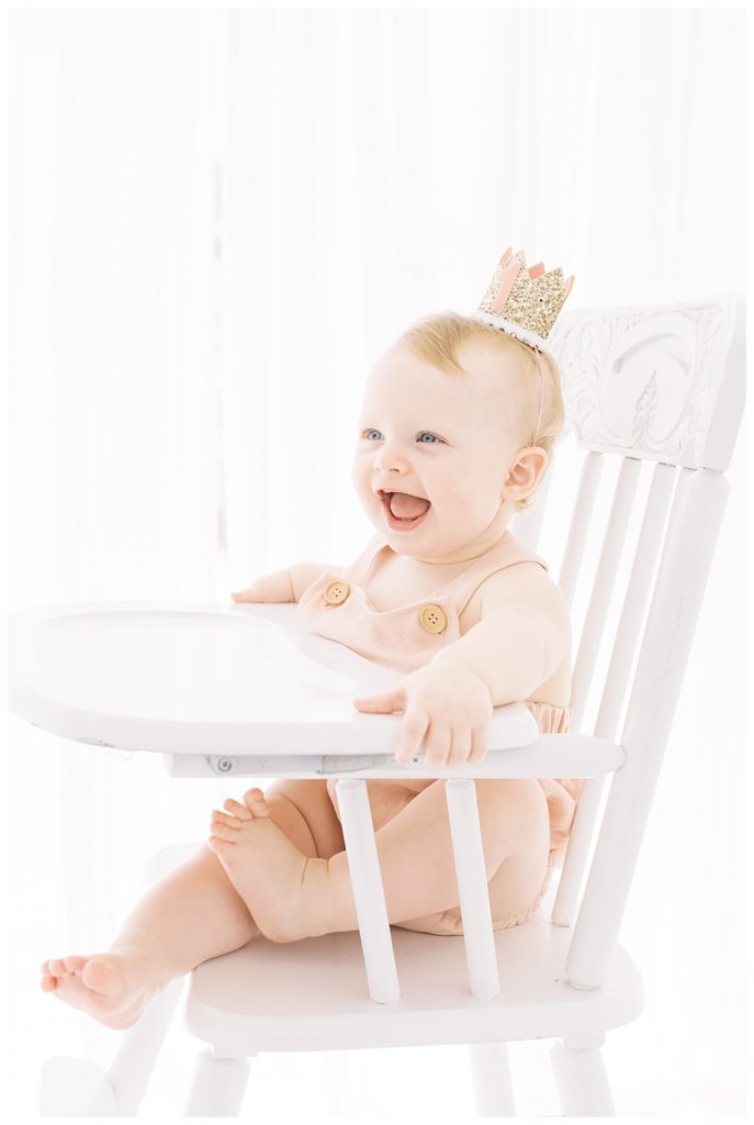 Baby Girl sits at high chair with a sparkly first birthday crown. Photographed by Halleigh Hill